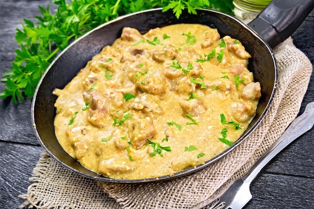 Carne cozida com creme em uma frigideira velha na serapilheira, salsa, garfo em um fundo de prancha de madeira