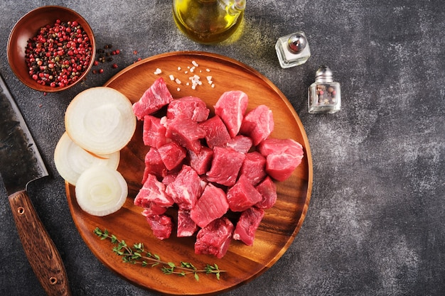 Foto carne cruda cortada en cubitos sobre una tabla de madera, especias, hierbas y verduras sobre fondo gris oscuro. ingredientes crudos para gulash. vista superior.