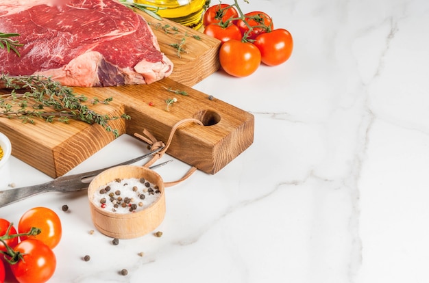 Carne cruda fresca, filete de mármol de cordero en una tabla de cortar, con ingredientes para cocinar. En la mesa de mármol blanco, copyspace