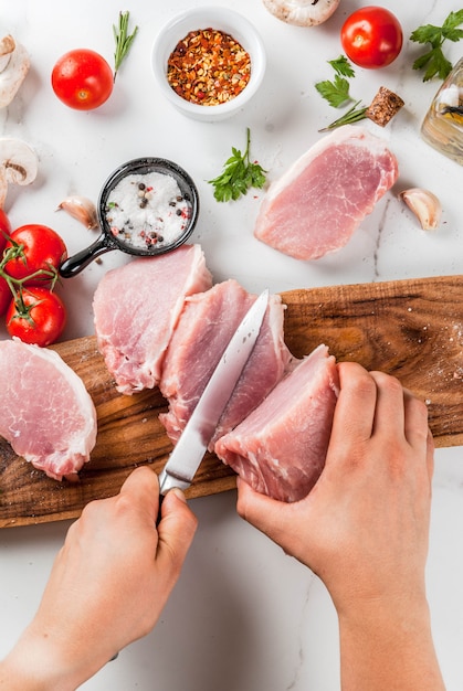 Carne cruda con ingredientes para la cena. Persona cortar filete de cerdo, lomo, en tabla de cortar, con sal, pimienta, perejil, romero, aceite, ajo, tomate, champiñones. Mesa de piedra negra, espacio de copia