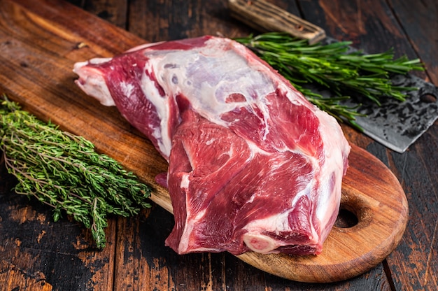 Carne cruda de paletilla de cordero o cabra en el hueso sobre una tabla de cortar de madera con cuchillo de carnicero. Fondo de madera oscura. Vista superior.