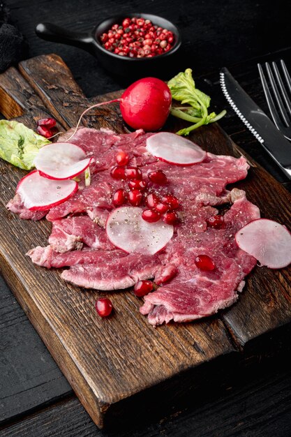 Carne de carpaccio com rabanete e granada na tábua de servir de madeira no fundo da mesa de madeira preta