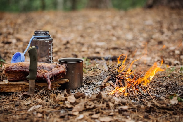 Carne de coelho grelhada na placa de madeira com garrafa de água de faca e caneca de titânio perto do fogo ao ar livre