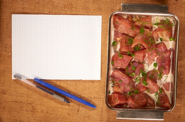 Carne marinada en leche y verduras en un plato hondo de acero sobre una tela casera con una hoja de papel limpia y un bolígrafo