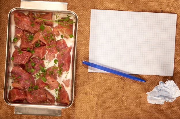 Carne marinada en leche y verduras en un plato hondo de acero sobre una tela casera con hojas de papel limpias y un bolígrafo