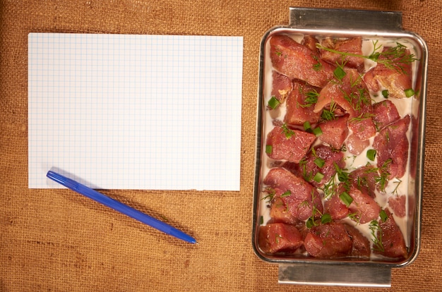 Foto carne marinada no leite e verduras em um prato fundo de aço em um pano caseiro com uma folha de papel limpa e uma caneta esferográfica
