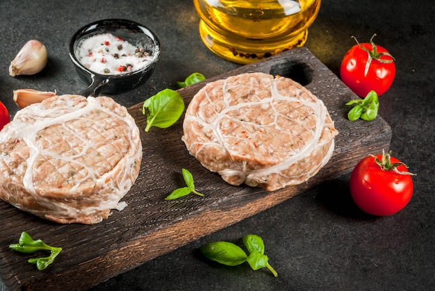 Carne orgánica cruda. Chuletas de pollo para hamburguesas, con parrilla de cerdo, para asar o freír. Con especias, albahaca, tomates, sobre una mesa de piedra gris sobre una tabla para cortar madera.