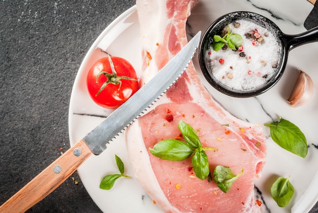 Carne orgánica cruda. Cocinar alimentos. Cerdo sobre hueso. Chuleta para freír a la parrilla. Con especias e ingredientes albahaca, ajo, sobre una mesa negra sobre una tabla de cortar de mármol blanco