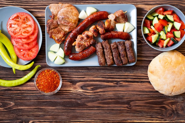 Carne a la parrilla tradicional serbia y balcánica llamada mesano meso. Barbacoa de los Balcanes (rostilj) servida con ensalada serbia, pimientos picantes, pan, tomate, cebolla y pimentón en polvo. Fondo de madera Vista superior