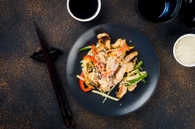 Carne de pollo al wok de verduras, salsas de soja y sésamo en un plato negro con palillos chinos sobre fondo oscuro. Tradición comida asiática. Copia espacio