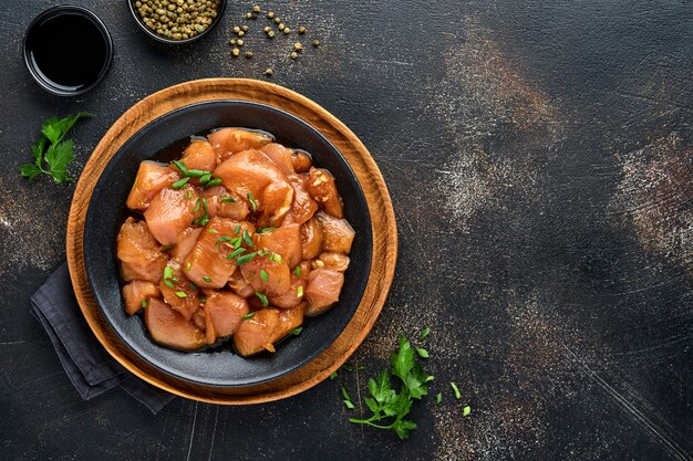 Carne de pollo cruda marinada en salsa de soja teriyaki, cebolla y pimiento en un plato negro sobre un fondo oscuro de pizarra, piedra u hormigón. Vista superior con espacio de copia.