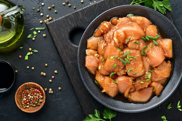 Carne de pollo cruda marinada en salsa de soja teriyaki, cebolla y pimiento en un plato negro sobre un fondo oscuro de pizarra, piedra u hormigón. Vista superior con espacio de copia.