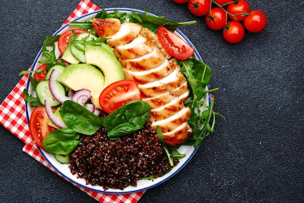 Carne de pollo a la parrilla, quinua roja y ensalada de verduras frescas con tomate, aguacate, rúcula y espinacas. Comida de dieta cetogénica de desintoxicación saludable. Vista superior de fondo negro.