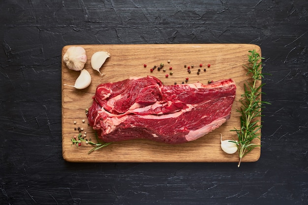 Carne de res cruda fresca con ajo y Romero en tabla de cortar sobre mesa de madera negra. Copie el espacio, arriba