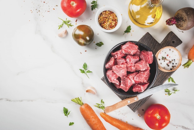 Carne de res. Goulash picado crudo fresco, cubos de carne en un tazón. Especias (sal, pimienta), tomate, ajo, cebolla. Sobre una mesa de mármol blanco, con un tenedor para carne y un cuchillo. vista superior