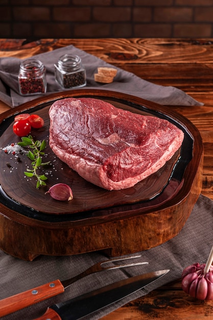 Carne de res de grupa de tapa cruda (picanha brasileña) sobre una tabla de cortar de madera resinada con especias. Mesa de madera con fondo de pared de ladrillos.