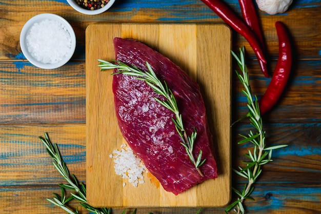 Carne de res orgánica cruda con condimentos de romero, sal y pimiento rojo sobre fondo de madera