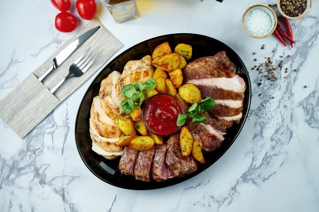 Carnes grelhadas variadas guarnecidas com batatas ao molho vermelho, servidas em prato preto sobre superfície de mármore