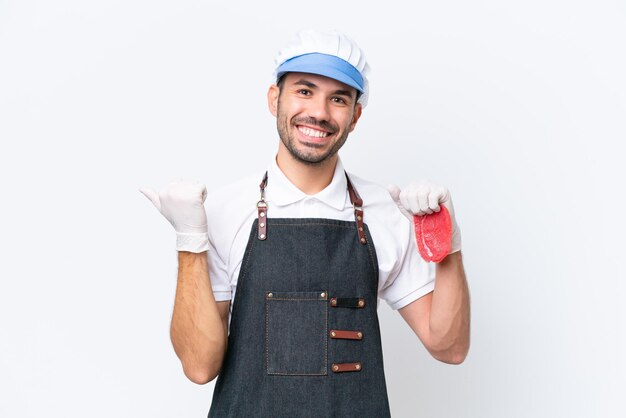 Carnicero hombre caucásico con un delantal y sirviendo carne fresca cortada sobre fondo blanco aislado apuntando hacia un lado para presentar un producto