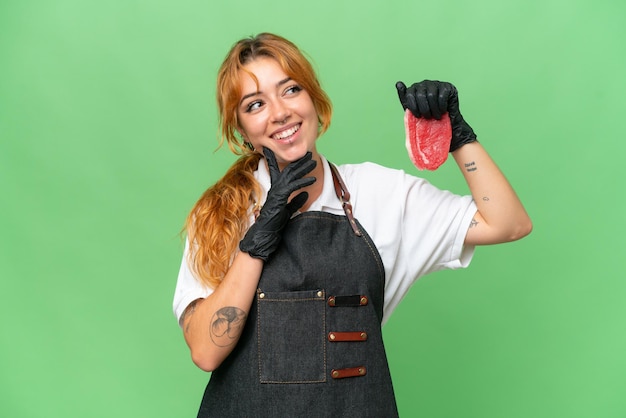 Carnicero mujer caucásica con un delantal y sirviendo carne fresca cortada aislada en el fondo de la clave de croma de pantalla verde pensando en una idea mientras mira hacia arriba