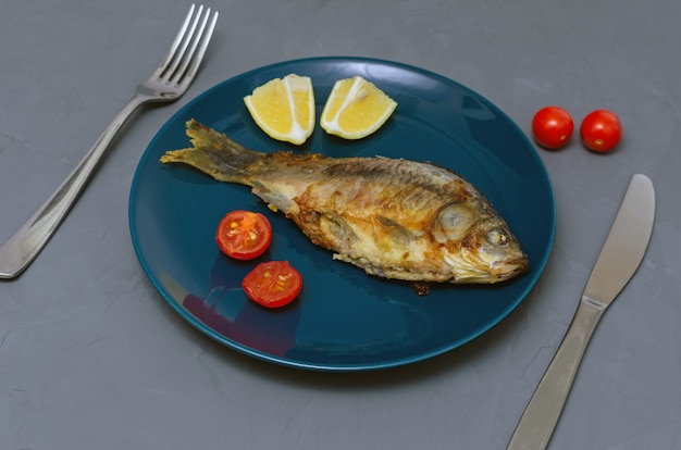 Carpa crucian de peixe apetitoso frito com tempero em um prato azul em uma mesa cinza decorada com fatias de tomate e limão com uma faca e um garfo. conceito de alimentação saudável.