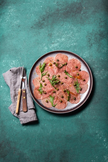 Carpaccio de carne marmoreada com rúcula e alcaparras na placa cinza vista superior de fundo azul