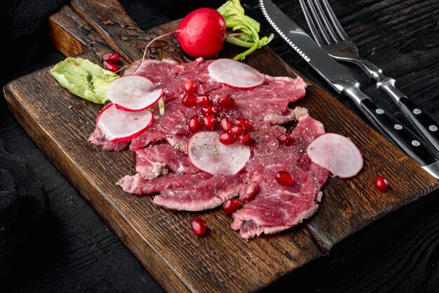 Carpaccio de carne marmorizada, com rabanete e granada, em tábua de madeira, sobre fundo de mesa de madeira preta