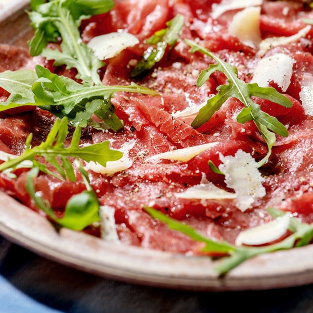 Carpaccio de ternera clásico con queso y rúcula en plato de cerámica sobre mantel blanco y azul. Imagen cuadrada