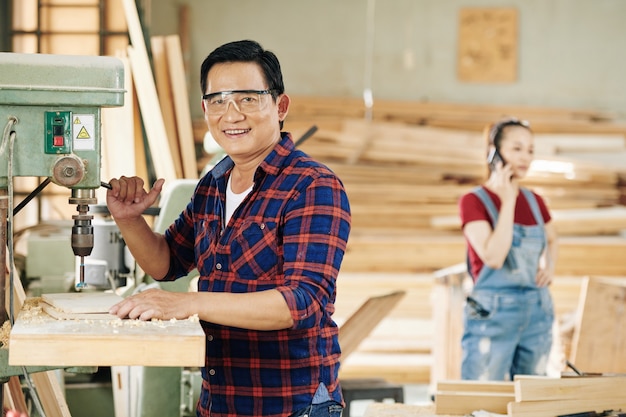 Carpinteiro asiático maduro em óculos de proteção fazendo furos em pranchas de madeira com a ajuda de uma máquina especial
