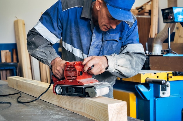 Carpinteiro em roupas de trabalho processa madeira com moedor Polimento da superfície da madeira Homem de meia idade trabalha na bancada na oficina de carpintaria Fluxo de trabalho real