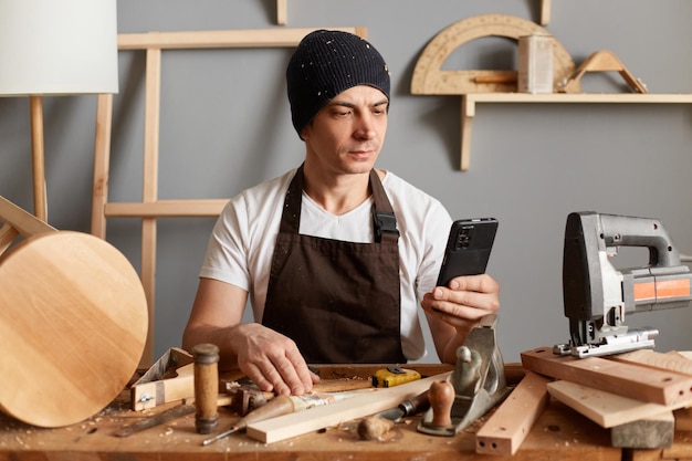 Carpinteiro jovem adulto vestindo um chapéu preto e avental marrom sentado à mesa de sua carpintaria usando seu telefone para procurar novas informações de carpintaria na internet usando seu telefone inteligente