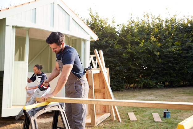 Carpinteiro masculino com aprendiz feminina serrando madeira para construir casa de verão ao ar livre no jardim