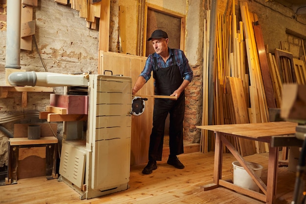 Carpinteiro sênior de uniforme trabalha com madeira em uma máquina de carpintaria na fabricação de carpintaria