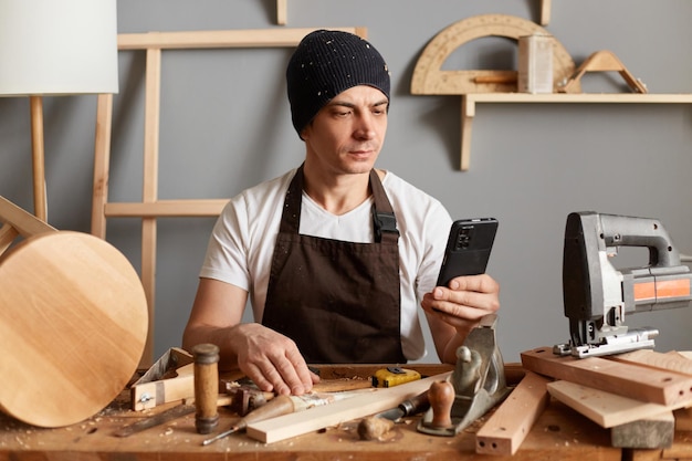 Carpintero adulto joven con un sombrero negro y un delantal marrón sentado en la mesa de su taller de carpintería usando su teléfono para buscar nueva información de carpintería en Internet usando su teléfono inteligente