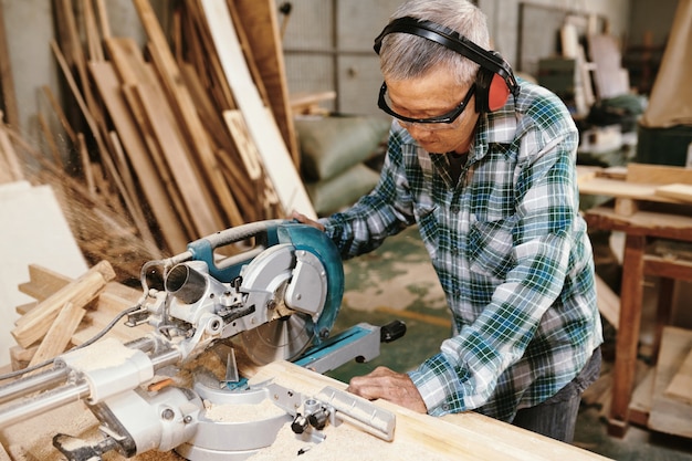 Carpintero anciano experimentado en gafas protectoras y orejeras cortando tablones de madera con sierra circular