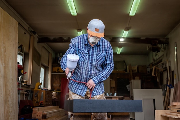 Carpintero experimentado en ropa de trabajo y propietario de una pequeña empresa está tallando una tabla de madera en un taladro de mano moderno en una vista lateral de taller ligero en el fondo muchas herramientas