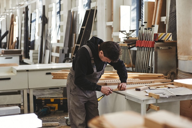 Carpintero experto en uniforme gris midiendo un trozo de madera en su taller de carpintería