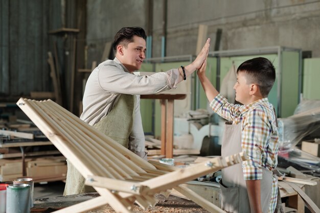 Carpintero masculino satisfecho en delantal dando cinco al hijo adolescente cuando han terminado la silla de madera en el taller