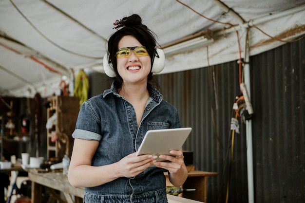 Carpintero mujer alegre con auriculares protectores usando una tableta en un taller