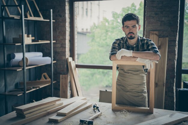 Carpintero serio confiado en guantes uniforme apoyado en bastidor artesanal mirándote pensativamente con tablas y herramientas en la mesa de lienzo