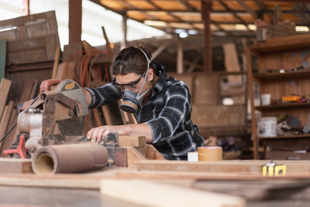 Carpintero de sexo masculino usa una máscara para evitar que las astillas de madera entren en su rostro mientras usa una máquina de sierra circular para cortar madera en un taller de carpintería.