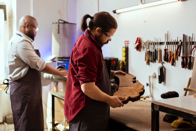 Carpintero sosteniendo un bloque de madera con un compañero de trabajo en el fondo iniciando la maquinaria
