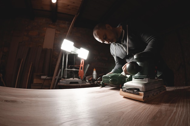 Un carpintero trabaja en un taller de fabricación de muebles de amoladoras de carpintería Un carpintero está amolando una pieza de madera con una lijadora eléctrica