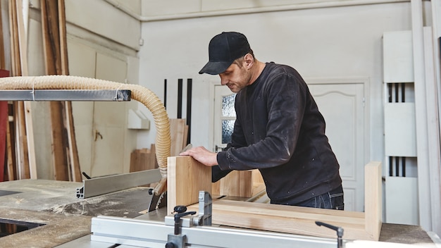 Carpintero trabajando en una sierra circular eléctrica cortando algunas tablas que lleva uniforme negro y una gorra