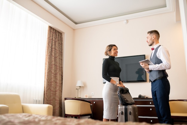 Carregador jovem e elegante com touchpad conversando com uma linda mulher de negócios com bagagem, ambas em pé no quarto de hotel