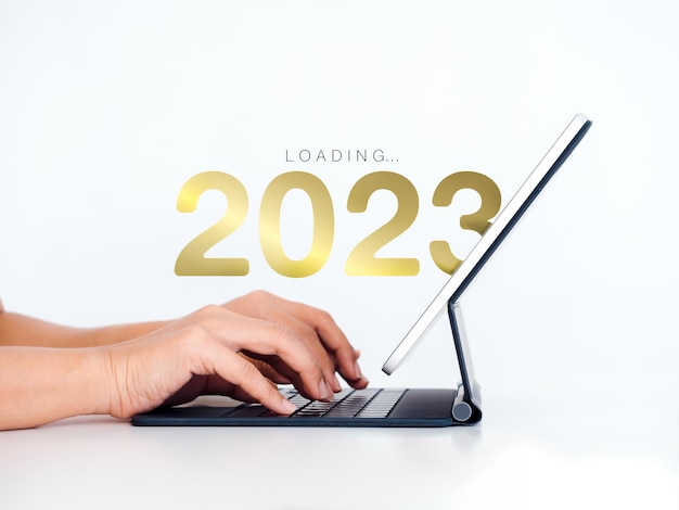 Carregando para 2023 feliz ano novo calendário de números do ano 2023 aparecem no computador tablet digital com as mãos digitando em fundo branco para se preparar para a mudança de ano novo e iniciar uma nova tecnologia de negócios