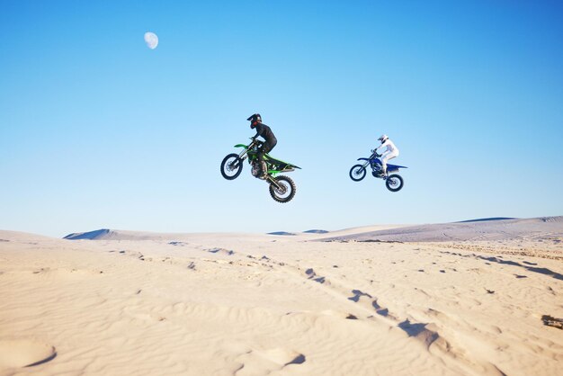 Carrera de motos en el desierto y salto en el aire para acrobacias de competencia y al aire libre para objetivos de rendimiento y velocidad Dunas de atletas de motos y rampa en la naturaleza rápido o juntos para concurso por el fondo del cielo