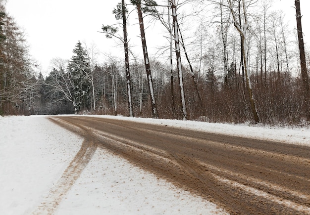 Carretera asfaltada en coche, donde la nieve se derritió. En la nieve se ven huellas de las ruedas del coche. Al costado de la carretera hay un bosque en crecimiento.