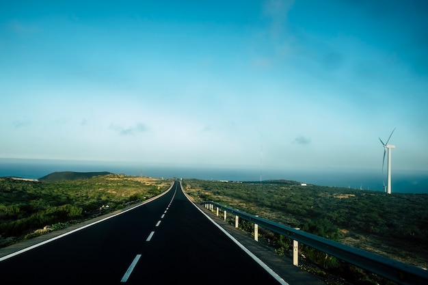 Carretera larga negra con franjas blancas en el medio para ir al océano. molino de viento en el lado derecho y naturaleza asombrosa alrededor. viajar y descubrir el concepto en un mundo hermoso