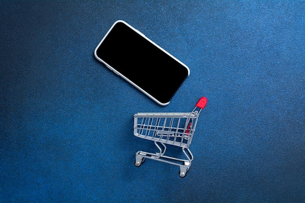 Carrinho de compras vazio e celular em um fundo azul escuro. Cesta de compras para produtos. Conceito de compras online